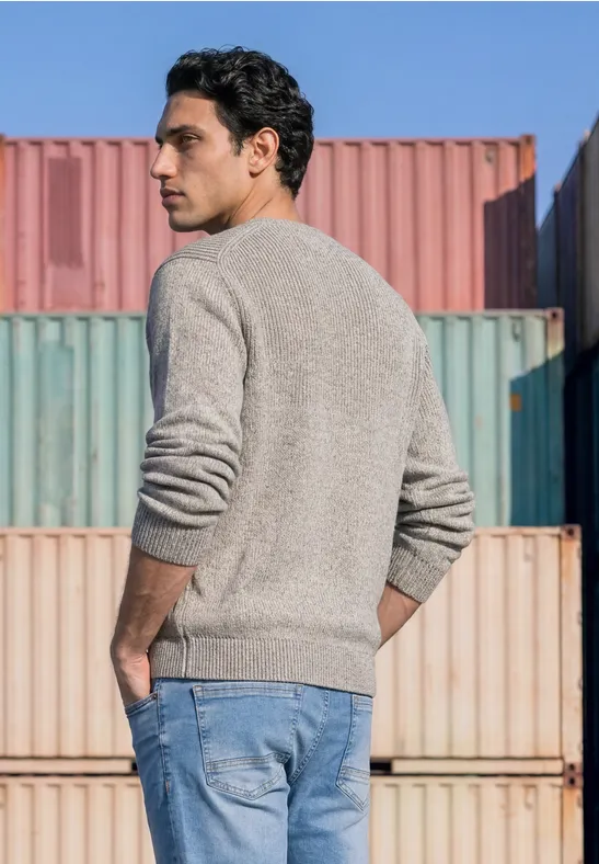Een man in een grijze trui en spijkerbroek staat voor gestapelde containers en kijkt opzij.