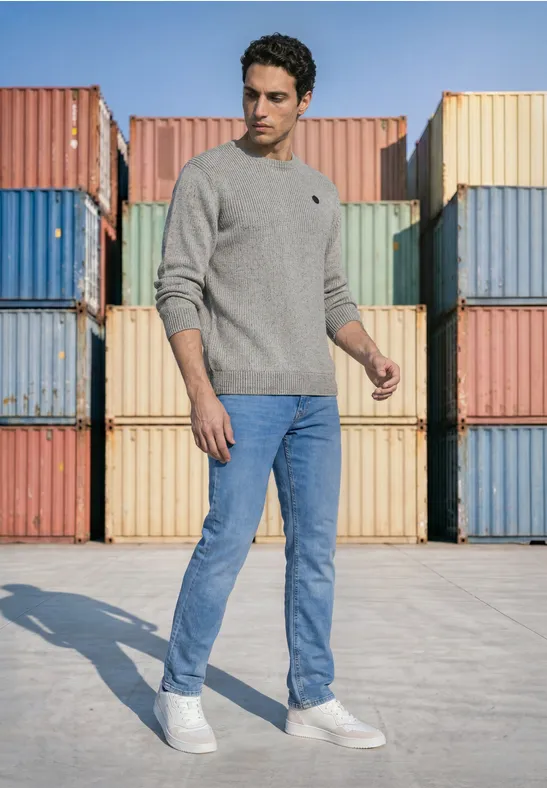 Man in een grijze trui, blauwe jeans en witte sneakers voor kleurrijke zeecontainers.