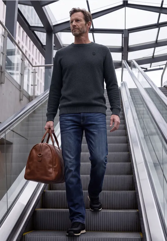 Een man in jeans en trui draagt een bruine leren tas op een roltrap in een modern, glazen gebouw.