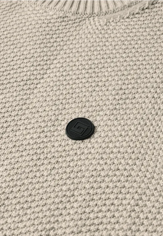 Beige trui met wafelgebreid patroon en een klein, rond zwart merklogo in het midden.