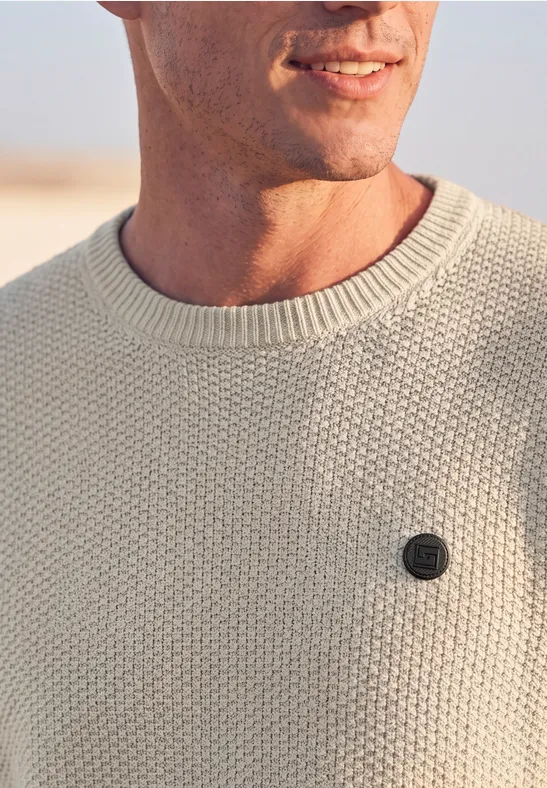 Close-up van een man in een cremekleurige gebreide trui met een klein rond embleem op de borst.