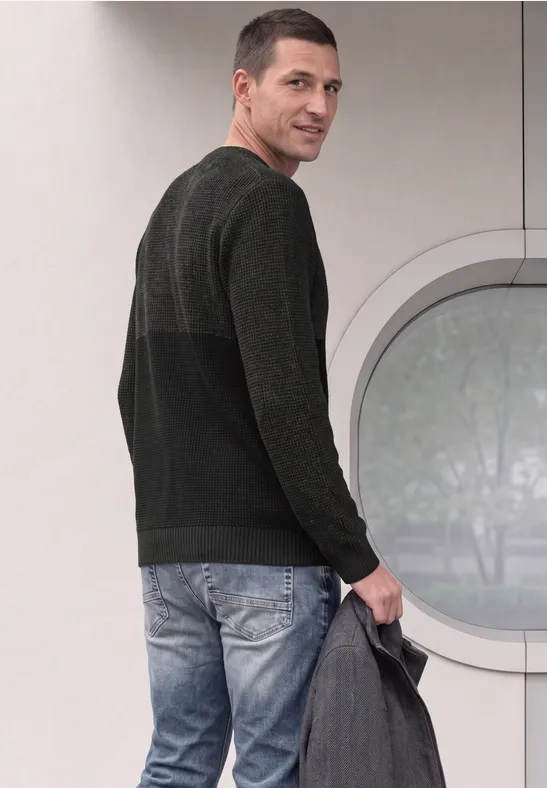Een man in spijkerbroek en donkere trui kijkt over zijn schouder en houdt een grijze jas vast.