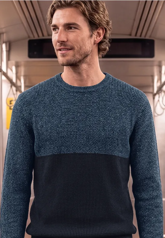 Een man in een tweekleurige trui staat in een metro- of treinwagon.