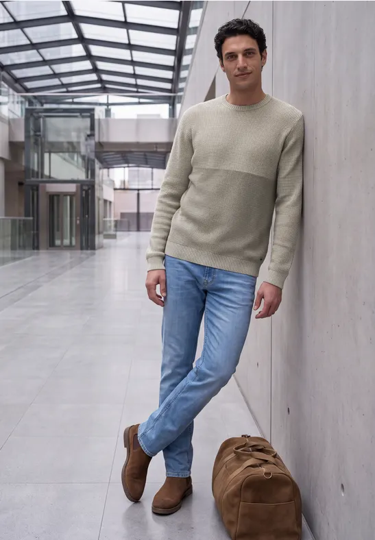 Een nonchalant geklede man leunt tegen een muur in een licht atrium, naast hem staat een grote reistas.
