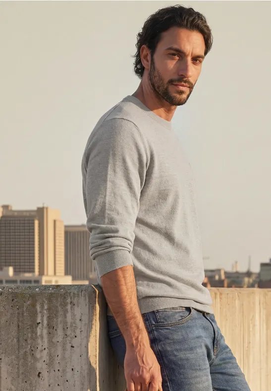 Ein lässig gekleideter Mann lehnt sich vor einer Betonmauer ab, trägt grauen Pullover und Jeans.