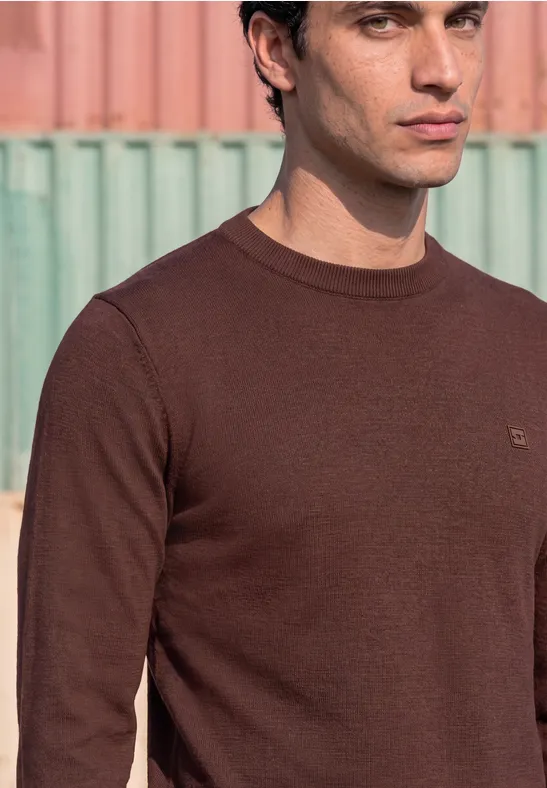 Ein Mann mit braunem Pullover vor pastellfarbener Wand.