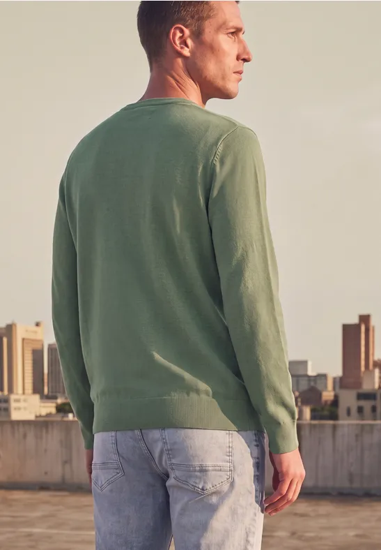 Ein Mann steht auf einem Dach, trägt grünen Pullover und Jeans, Blick zur Stadt im Abendlicht.