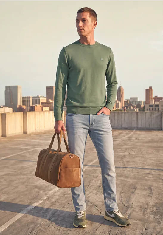 Mann auf Parkhausdach mit grünem Pullover, blauen Jeans, grünen Sneakern und brauner Reisetasche vor Stadtkulisse.