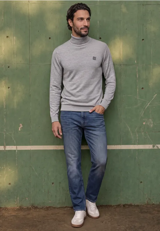 Man in grijze coltrui, blauwe jeans en witte sneakers voor een groene houten achtergrond.