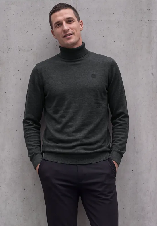 Een man staat nonchalant met handen in zakken tegen een grijze muur, gekleed in een donker coltrui.