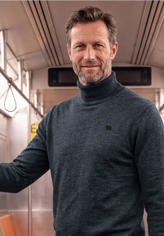 Een man in een grijze coltrui glimlacht in de metro.