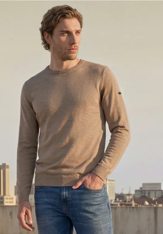 Een man in een bruin trui en spijkerbroek staat op een dak met een stedelijke skyline op de achtergrond.