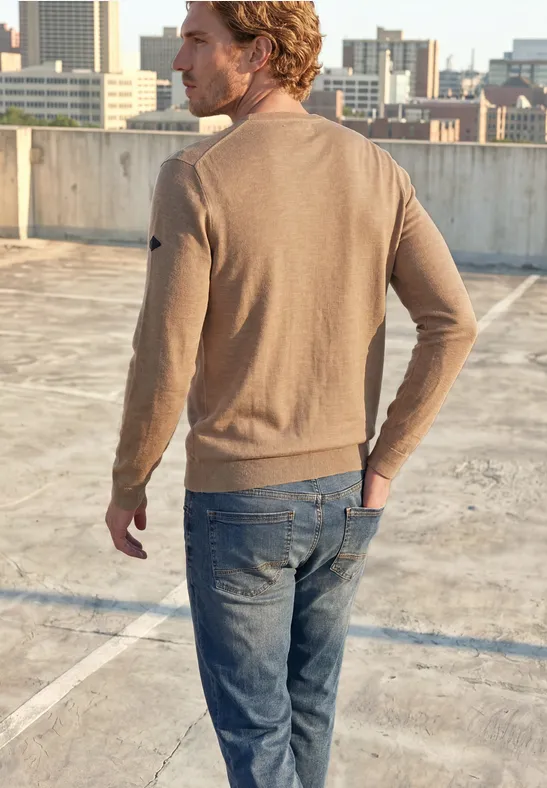 Een man in een bruine trui en spijkerbroek staat op een parkeerdak, uitkijkend over de stad.