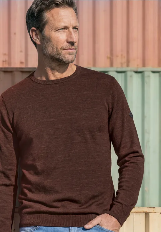 Ein Mann in braunem Pullover steht vor Containern und blickt zur Seite.