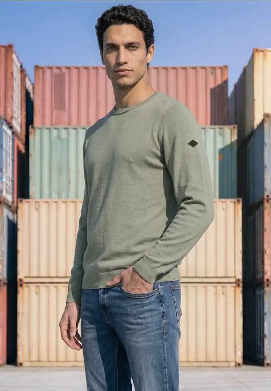 Ein Mann in grünem Pullover und Jeans steht vor bunten Frachtcontainern.