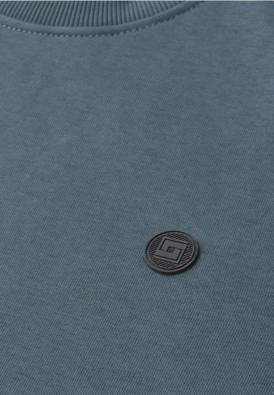Nahaufnahme eines blauen Sweatshirts mit rundem schwarzen Logo-Patch neben dem Kragen.