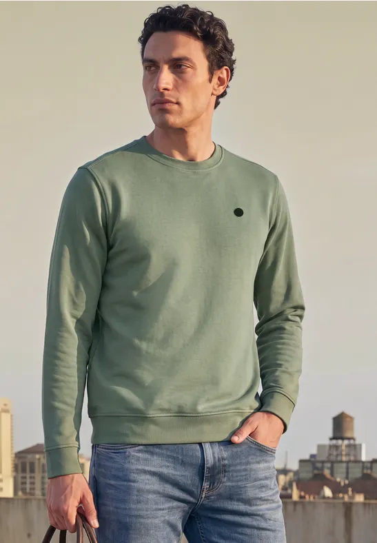 Een man draagt een groen sweatshirt en jeans, staand voor een stad bij zonsondergang.