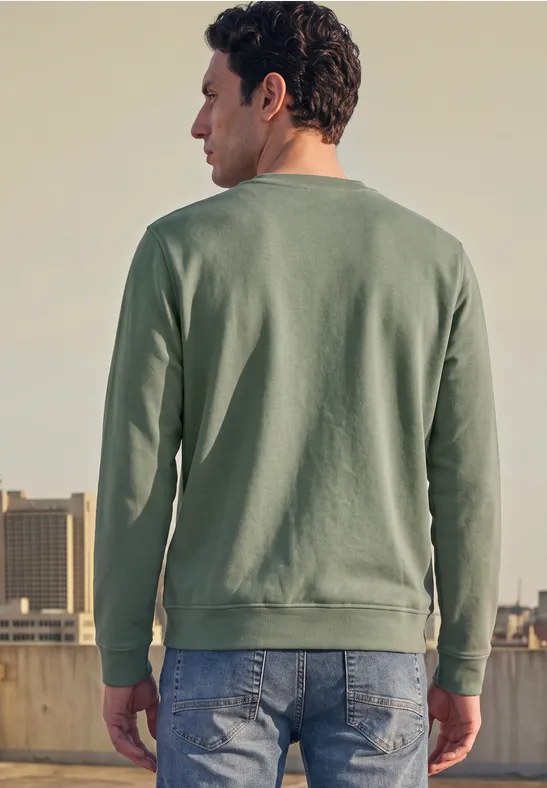 Een man staat op een dak, rug naar de camera, draagt een groen sweat en jeans voor een stadsachtergrond.
