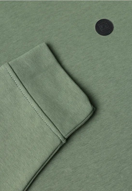 Close-up van een groen kledingstuk met zak en rond zwart logo.