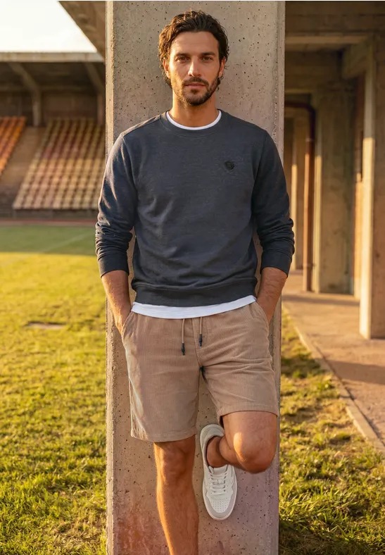 Mann lehnt lässig an Säule im Freien, trägt Sweatshirt, Shorts und weiße Sneaker.