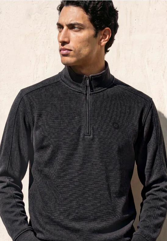 Ein Mann mit dunklem, schmal geschnittenem Zip-Pullover steht vor hellem Hintergrund.