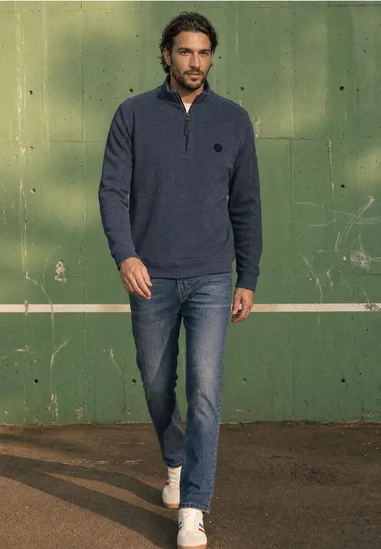 Ein Mann in Jeans, weißen Turnschuhen und blauem Zip-Pullover geht vor einer grünen Wand mit weißer Linie.