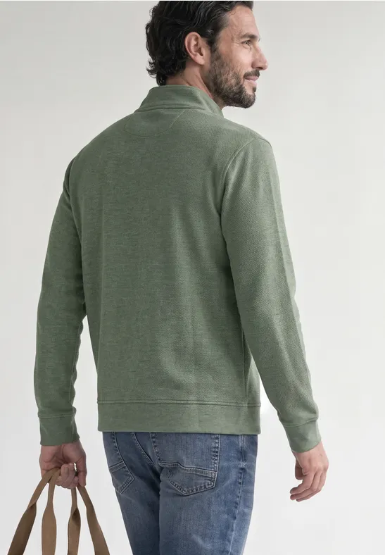 Ein Mann mit grünem Pullover und Jeans, Rückenansicht, hält eine Tasche in der Hand.