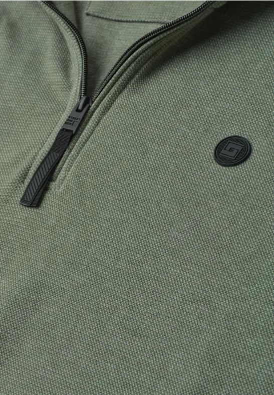 Nahaufnahme eines olivgrünen Strickpullovers mit halbem Reißverschluss und rundem Logo-Patch.