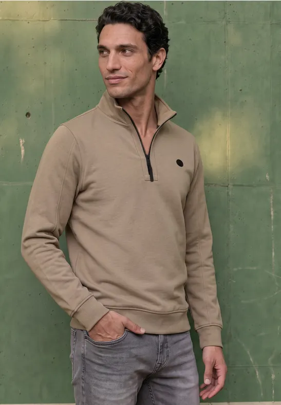 Ein Mann steht vor einer grünen Wand, trägt einen beigen Pullover mit kurzem Reißverschluss und Jeans.