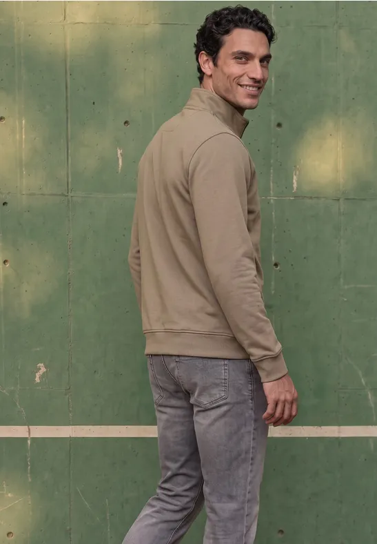 Ein Mann steht seitlich vor grüner Wand und schaut über die Schulter, trägt olivgrünen Pullover und graue Jeans.