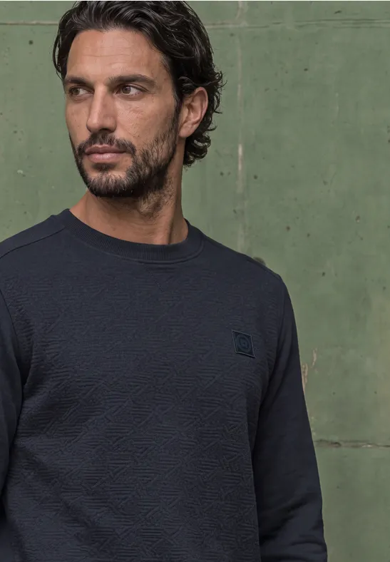Ein Mann steht in dunklem Pullover vor einer grünen Wand und schaut nach rechts.