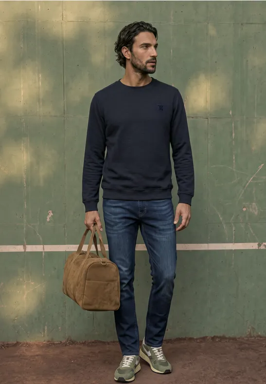 Ein Mann in dunkelblauem Pullover, Jeans und Sneakers steht vor grüner Wand und hält eine braune Tasche.