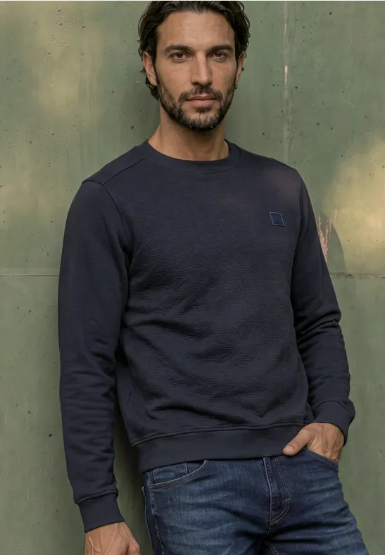 Ein Mann steht vor einer grünen Wand, trägt einen dunkelblauen Pullover und blaue Jeans.