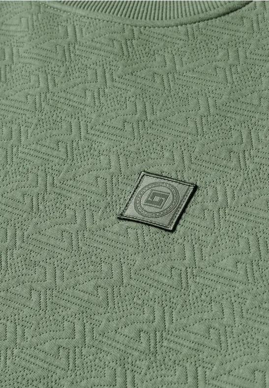 Zielona tkanina z geometrycznym wzorem na całej powierzchni i małą czarną kwadratową naszywką z logo.