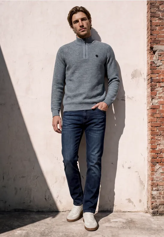 Een man leunt ontspannen tegen een zonnige muur, draagt een grijze trui, blauwe jeans en lichte sneakers.