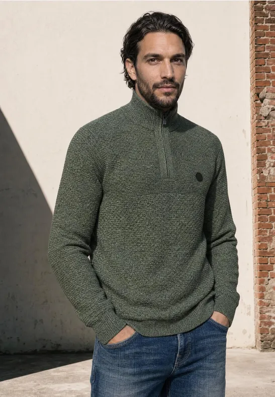 Ein Mann trägt einen grünen Pullover mit kurzem Reißverschluss und Jeans und steht vor einer Wand.