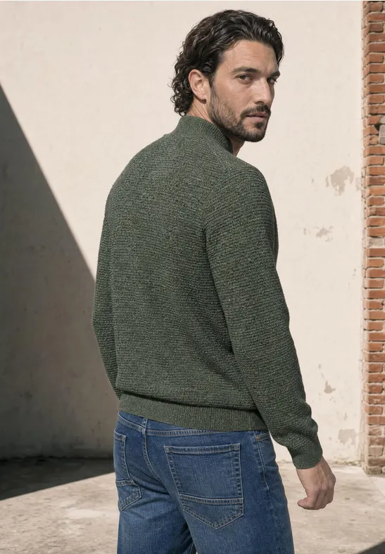 Ein Mann im grünen Pullover und Jeans steht draußen und blickt über die Schulter.