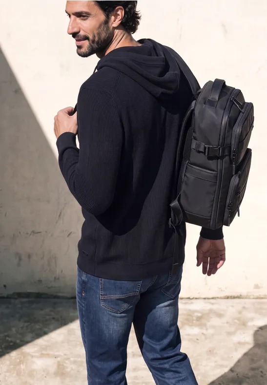 Un homme en sweat à capuche noir et jean porte un sac à dos noir en marchant devant un mur clair.