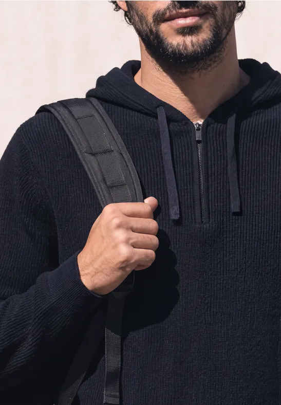 Un homme en sweat à capuche bleu foncé tient la bretelle de son sac à dos sur l’épaule