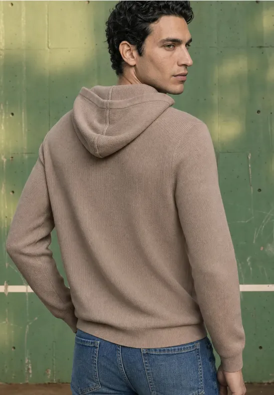 Een man van achteren in een beige hoodie met spijkerbroek voor een groene achtergrond.