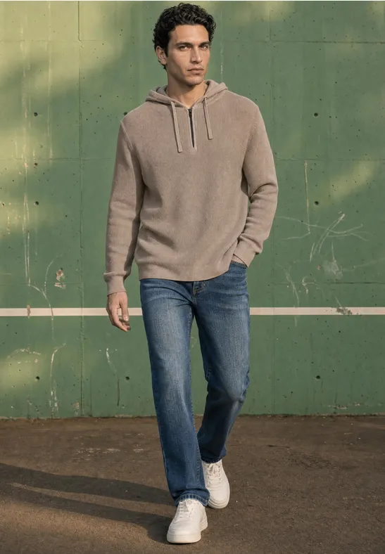 Een man in een beige hoodie, spijkerbroek en witte sneakers loopt voor een groene muur.