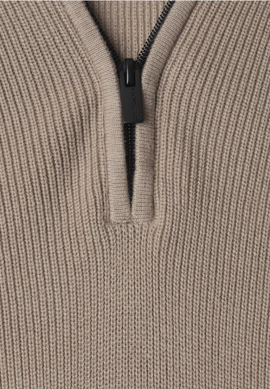 Close-up van een beige grofgebreide trui met korte rits en opstaande kraag.