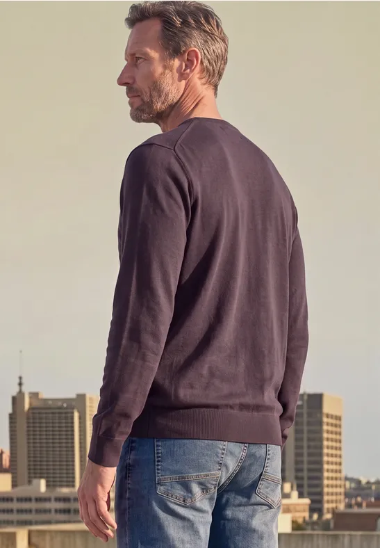 Un homme en pull et jean, vu de dos, se tient devant un décor urbain.