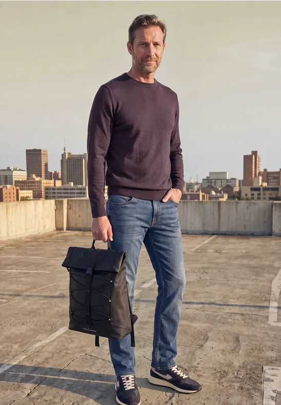 Un homme est sur un toit-terrasse, vêtu d'un pull, jean et baskets, tenant un sac à dos noir.