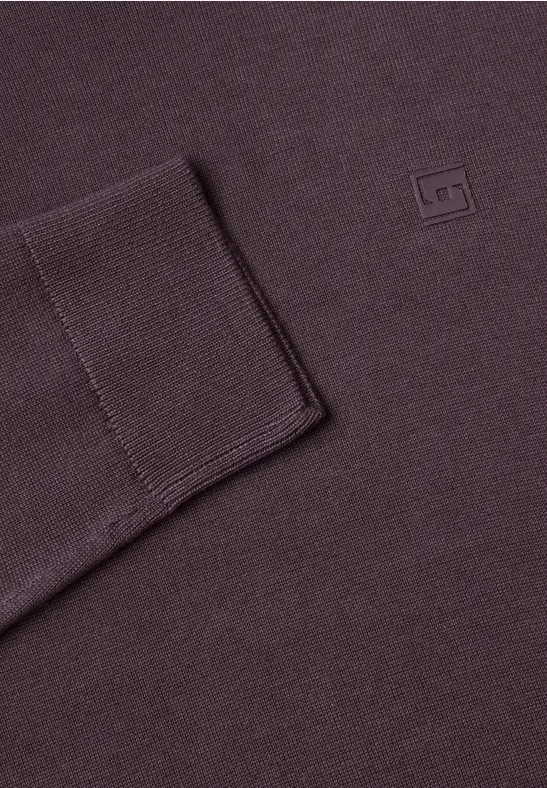 Gros plan sur un pull tricoté aubergine, avec poignets côtelés et petit logo carré.