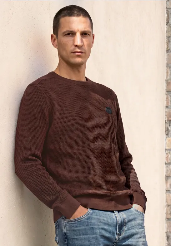 Ein Mann lehnt an einer Wand, trägt einen braunen Pullover und Jeans.