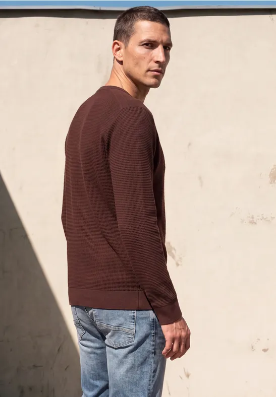 Ein Mann steht vor einer hellen Wand, trägt braunen Pullover und Jeans und blickt über die Schulter.