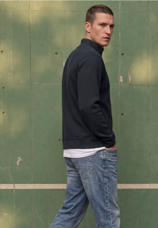 Un homme en jean et pull se tient devant un mur vert et regarde en arrière.