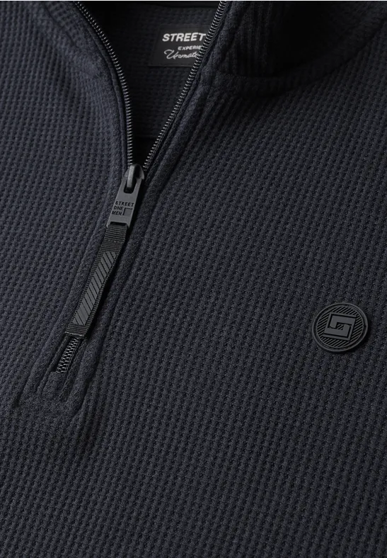 Gros plan d’un haut noir en maille à demi-zip et motif nid d’abeille, avec un petit logo discret.