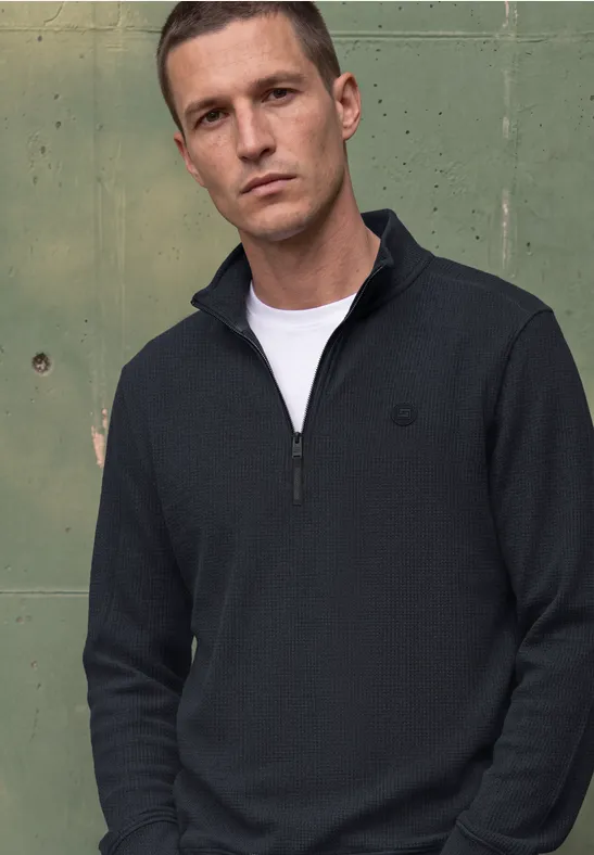 Un homme devant un mur métallique vert porte un pull bleu marine à demi-zip et un t-shirt blanc.